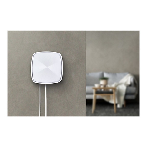 WeiÃŸes, quadratisches GerÃ¤t an grauer Wand, darunter zwei Kabel. Im Hintergrund verschwommenes Wohnzimmer.