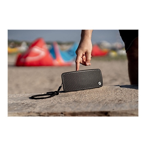 Ein Mann berührt mit dem Finger einen schwarzen Lautsprecher auf einem Stein. Im Hintergrund sind Strand und Kitesurfer.