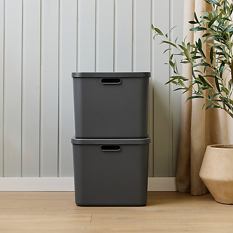 Zwei graue Aufbewahrungsboxen mit Deckel, übereinander. Holzfussboden und Wand mit Holzvertäfelung im Hintergrund, rechts grüne Pflanze.