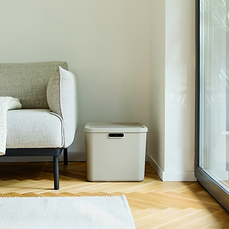 Hellgraue Eimer und Boxen stehen in einer Ecke vor einem Sofa. Der Hintergrund ist hell und die Szene wirkt ruhig.