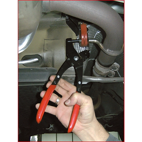 Hand hält Spezialzange am Auspuff. Schwarze Zange mit roten Griffen. Im Hintergrund Auspuff und Fahrzeugunterseite.