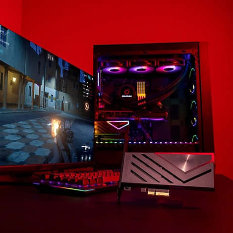 Eine AVerMedia Capture Card liegt auf einem Tisch vor einem leuchtenden Gaming-PC und einem Monitor.
