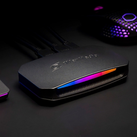 Schwarze AVerMedia Capture Card mit RGB-Beleuchtung auf einem dunklen Tisch, angeschlossene Kabel sichtbar.