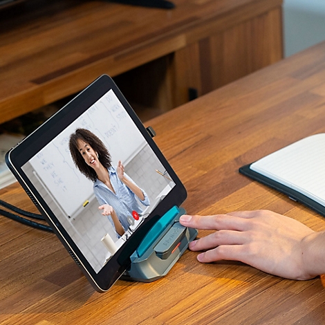 Ein Tablet in einem Standfuß auf einem Holztisch zeigt eine Frau bei einem Online-Unterricht per Videokonferenz.