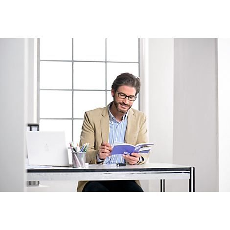 A man with glasses reads a blue book at a desk in a bright office. He is wearing a beige jacket.