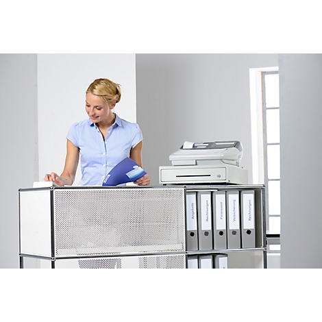 A smiling woman in a blue shirt stands at a desk with binders, documents, and a printer. She is looking at a blue document.