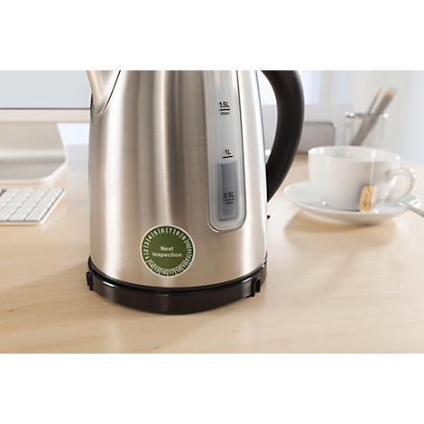 A stainless steel kettle sits on a table. Sticker "Next Inspection". A cup of tea next to it.