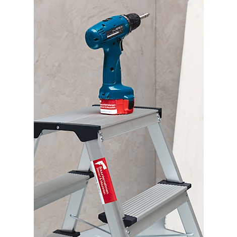A blue cordless drill stands on a small silver stepladder. Red battery, gray background. A red sticker is attached to the ladder.
