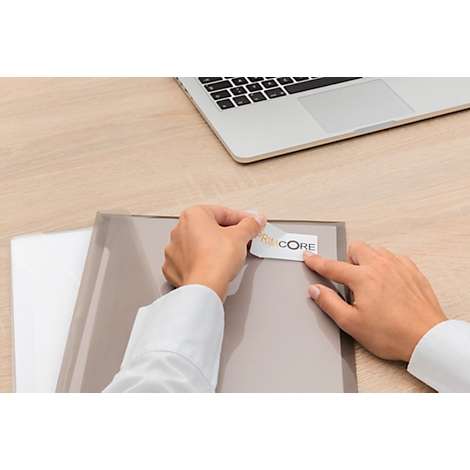 Person sticking a sticker labeled "CORE" onto a gray folder. A laptop rests on the table.