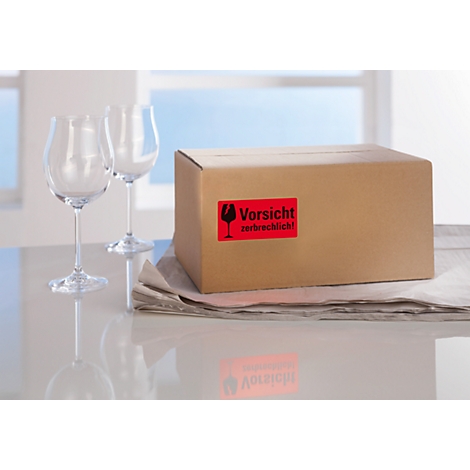 Two wine glasses stand next to a package with the red sticker "Fragile!" in German.