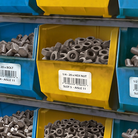 Gelbe und blaue Behälter mit Schrauben und Muttern in einem Lager. Beschriftungen mit Produktbezeichnungen und Barcodes.