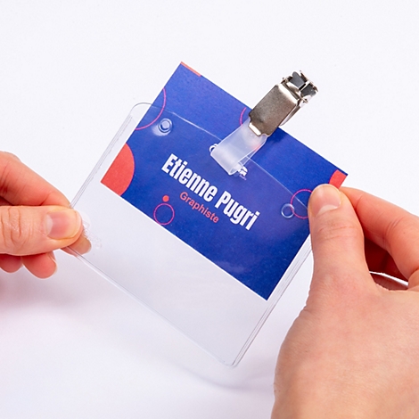 Handen houden een naambadge met een metalen clip en blauwe inlegvel met 'Etienne Pugri, Graphiste'.