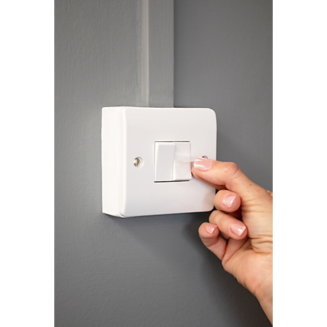 A finger presses a light switch on. The switch is fixed in a white holder on a gray wall.