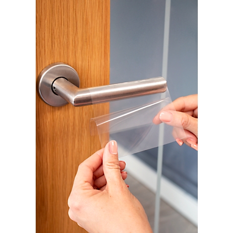 Hands holding a transparent protective film to cover a metal door handle. The door handle is attached to a wooden door.