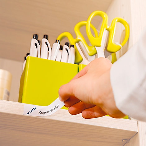 A hand sticking a label that says "Kugelschreiber" on a shelf containing pens and scissors.