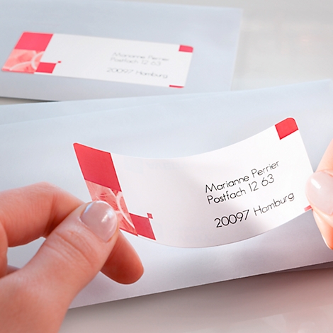Close-up of two hands attaching an address label with a red design to an envelope. The text on the label reads: Marianne Perrier, Postfach 12 63, 20097 Hamburg.