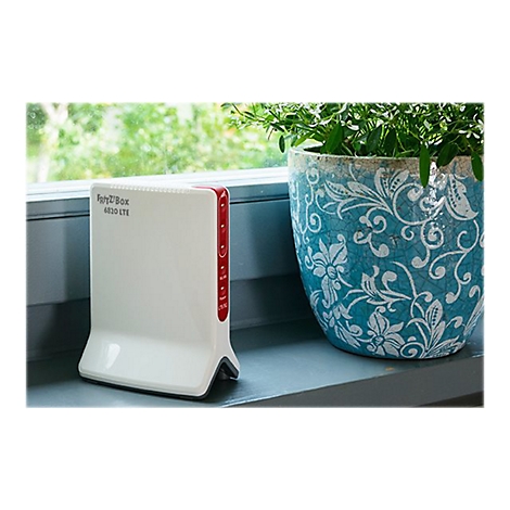 Fritz!Box 6820 LTE blanche sur un rebord de fenêtre. À droite, un pot avec des plantes vertes, derrière, un paysage vert flou.