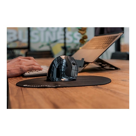 Ein ergonomische Maus liegt auf einem schwarzen Mauspad, daneben ein Laptop auf einem Ständer. Eine Hand bedient eine Tastatur.