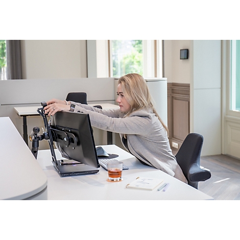 Eine blonde Frau im grauen Blazer streckt sich im Büro. Sie sitzt vor einem Bildschirm, Hände am Monitor.