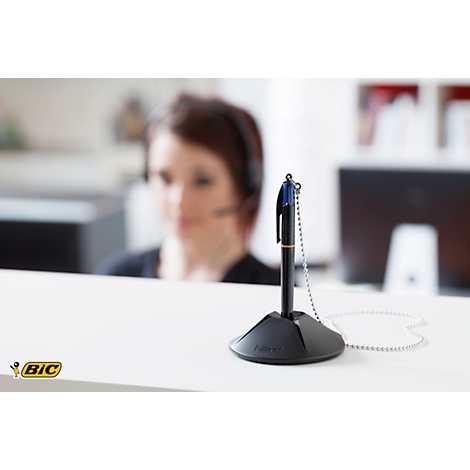 Pen with chain in a pen holder. In the background a woman with a headset at the desk.