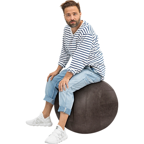 Un hombre sentado en un balón de ejercicio redondo y marrón. Lleva vaqueros, una camisa a rayas y zapatillas blancas.