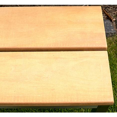 Gros plan sur un banc en bois brun clair avec des bords arrondis. En arrière-plan, on voit de l'herbe et un mur de pierre grise.
