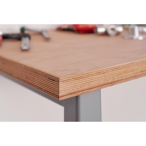 Primer plano de una mesa de madera con capas visibles, sobre una pata de mesa gris. Las herramientas están borrosas en el fondo.