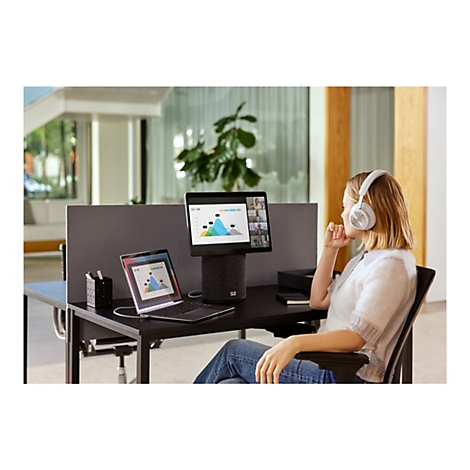 Eine Frau sitzt an einem Schreibtisch mit Headset und schaut auf ein Tablet mit Videokonferenz. Auf dem Tisch stehen ein Laptop und ein zylinderförmiger Lautsprecher.