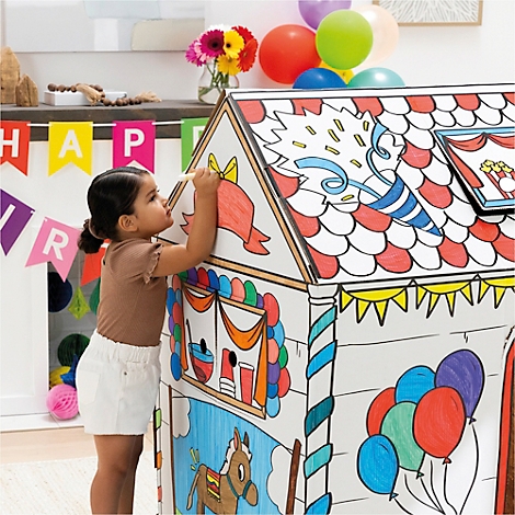 Une petite fille colorie une maison de jeu blanche avec des crayons, décorations de fête en arrière-plan.