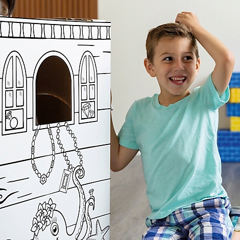 Een lachende jongen staat naast een zwart-wit geschilderde piratenkist met sieraden. Hij heeft een hand op zijn hoofd.