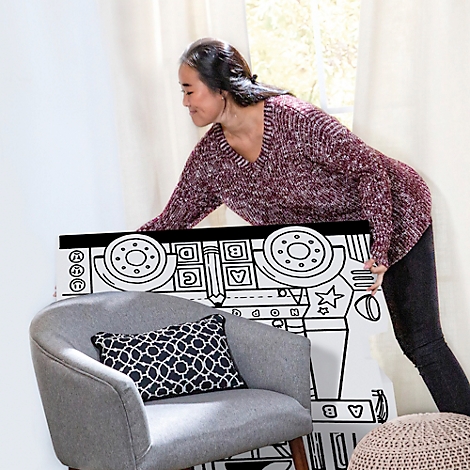 Een vrouw houdt een zwart-wit kleurplaat vast, die in een fauteuil staat. Een raam op de achtergrond.