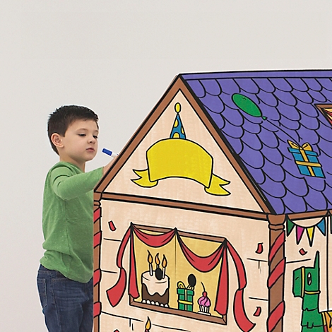 Een jongen met een groen shirt schildert een kartonnen huis. Op het dak staan versieringen zoals een ballon, een cadeau en een verjaardagskroon.