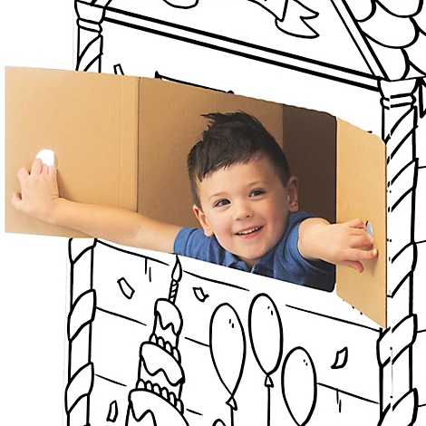 Een lachende jongen in een kartonnen huis, versierd met een verjaardagstaart en ballonnen, zwaait naar de camera.