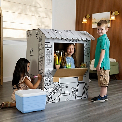 Trois enfants jouent avec un magasin en carton, l'un est dans la fenêtre, deux le peignent. Une glacière est à côté.