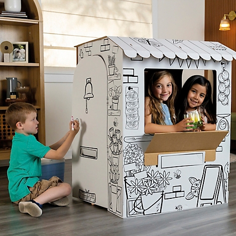 Un enfant dessine sur une boutique de jouets. Deux filles sont assises à la fenêtre et sourient. L'étal est dessiné sur du carton.