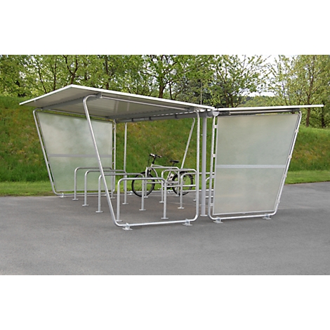 Bike shelter with bike. Metal canopy, transparent side walls. On asphalt, green trees in the background.