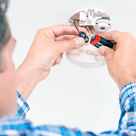 Man changing batteries in a smoke detector on the ceiling. He holds a new battery.