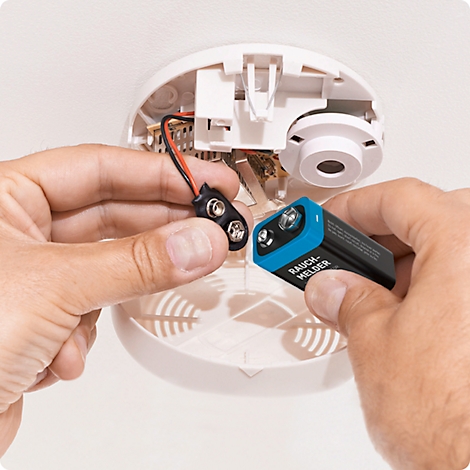 Hands are inserting a battery into a smoke detector. The smoke detector is mounted on the ceiling.