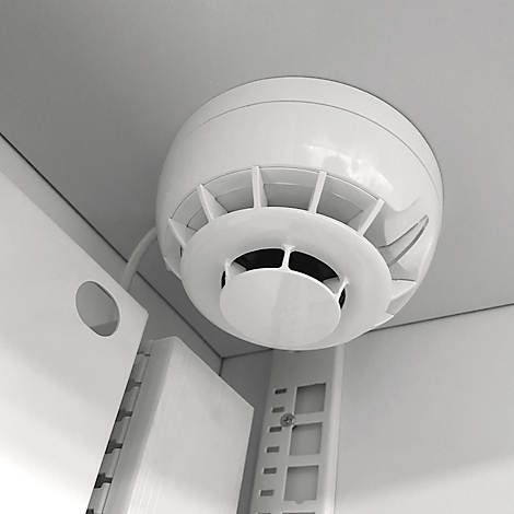 Weißer Rauchmelder an weißer Decke mit Lüftungsschlitzen. Kabel und Halterung sind ebenfalls zu sehen.