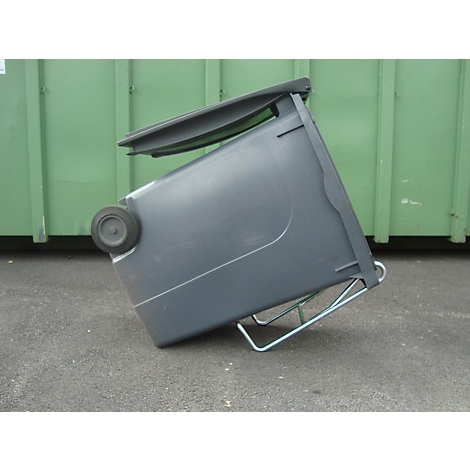 Ein grauer Mülleimer kippt auf einer befestigten Fläche. Im Hintergrund ein grüner Container.