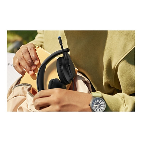 Nahaufnahme: Person hält schwarzes Headset mit Mikrofon. Person trägt grüne Uhr und Pullover. Gelber Notizblock.
