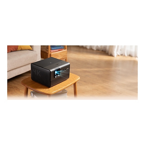 Ein schwarzer 4K-Beamer auf einem kleinen Holztisch. Das GerÃ¤t projiziert blaues Licht, im Hintergrund Sofa und Fenster.