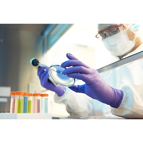 Laborant met bril en masker onderzoekt een petrischaal met een pipet. Reageerbuizen op de voorgrond.