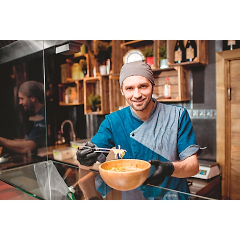 Een chef-kok lacht, terwijl hij een kom ramen vasthoudt met eetstokjes. Hij draagt een muts, een koksjas en handschoenen.