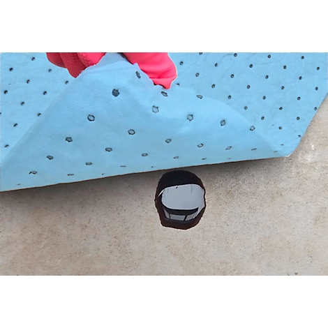 Helle, blaue Flüssigkeitsbindematte bedeckt dunkle Flüssigkeit auf beiger Oberfläche. Eine rosa-gehandschuhte Hand hebt die Matte an.