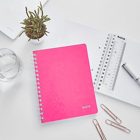 Un cahier rose à spirales sur une table blanche. A côté : règle, stylo, plante et trombones.