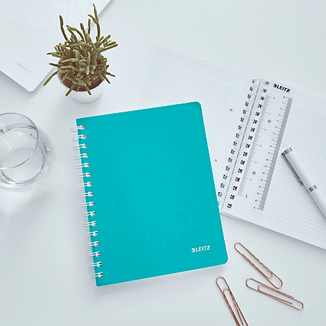 Cahier turquoise avec spirale, à côté règle, stylo et trombones sur surface blanche.