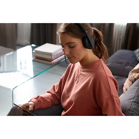 Woman with headphones focused on tablet, sitting on sofa. Background: couch, table and books.