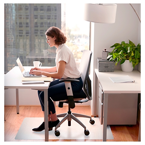 Frau sitzt an Schreibtisch und arbeitet am Laptop. Sie trägt ein weißes Hemd und eine dunkle Hose. Fenster mit Skyline.