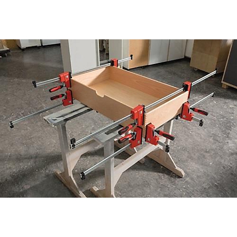 Wooden box fixed with clamps on workbench. Wooden box, red-black clamps, metal rods, light wood surface.
